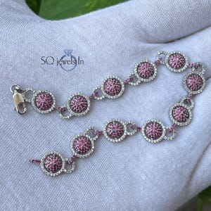 May include: A silver bracelet with a series of circular, pink gemstone clusters, each surrounded by a halo of smaller, clear stones. The bracelet is displayed on a white surface. The text "SQ jewels In" is visible.