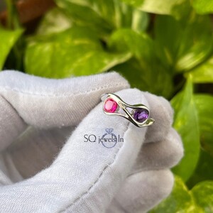 May include: A silver ring with a pink and purple gemstone design. The ring has an openwork design with a pink gemstone on one side and a purple gemstone on the other. The ring is set against a background of green foliage.