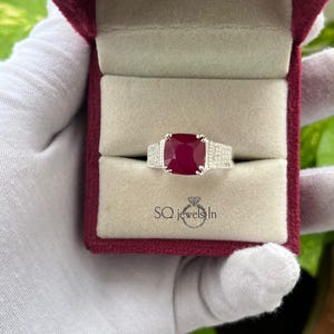 May include: A silver ring with a large, square, deep red gemstone, flanked by smaller, clear stones. The ring is displayed in a burgundy velvet ring box. The text "SQ jewels In" is visible.