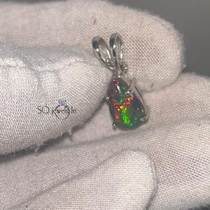 A 925 sterling silver pendant featuring a pear-shaped cabochon natural black Ethiopian opal with fiery green and red fire, held in a 3-prong setting with a polished bail over a gloved hand.