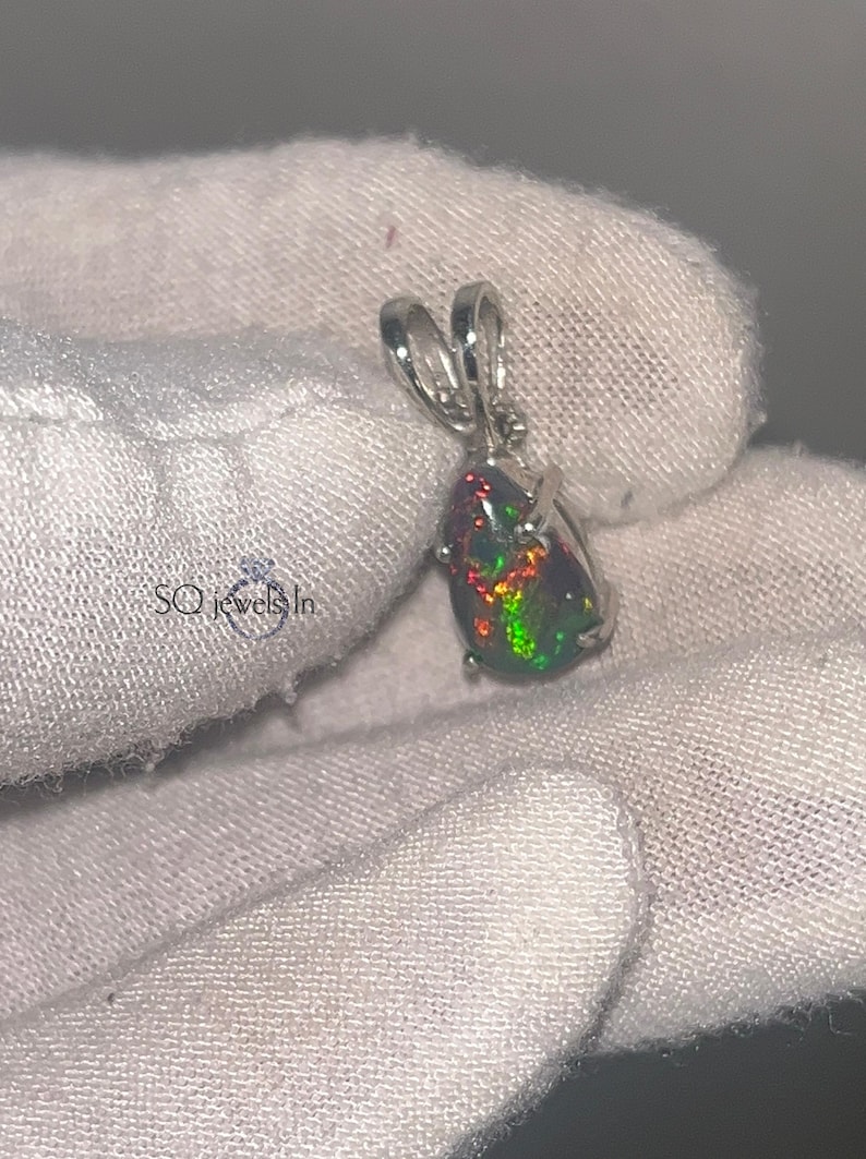 A 925 sterling silver pendant featuring a pear-shaped cabochon natural black Ethiopian opal with fiery green and red fire, held in a 3-prong setting with a polished bail over a gloved hand.