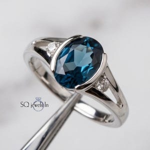 May include: A silver-toned ring featuring a large, oval-cut, deep blue gemstone set in a tension-style mount. The band includes two small, clear, round-cut accent stones on either side of the center gem. The text SQ jewels In is visible.