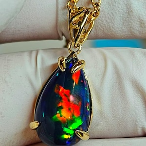 Può includere: Una collana con pendente in oro con un'opale nera a forma di goccia con vibranti colori interni rossi, arancioni e verdi. L'opale è incastonata in una lunetta d'oro e sospesa a una catena d'oro. Il timbro data e ora "27 luglio 2023 15:31" è visibile.