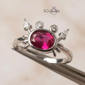May include: A silver-toned ring featuring a crab-shaped design with a large oval-cut vibrant pink center stone. The crab's eyes are set with two small round white stones, and its claws are accented with two pear-cut white stones. Text reads SQ Jewels In.