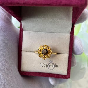 Puede incluir: Un anillo de oro con un diseño floral, con una piedra central marrón oscuro rodeada de piedras preciosas amarillas más pequeñas. El anillo se presenta en una caja de joyería burdeos con interior blanco. El texto "SQ jewels In" es visible.