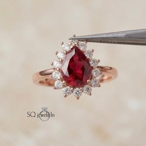 May include: Rose gold ring featuring a pear-shaped, deep red gemstone surrounded by a halo of smaller, white gemstones. The ring is held by tweezers against a soft, neutral background. The ring's band is a delicate rose gold.