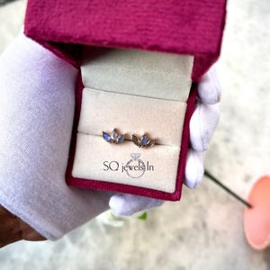 May include: A pair of gold-toned stud earrings with blue-toned gemstones, presented in a magenta velvet jewelry box. The box is open, revealing the earrings nestled in a cream-colored interior. The text "SQ jewels In" is printed on the box.