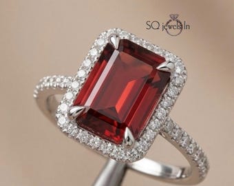 May include: A silver ring featuring a large, rectangular, deep red gemstone surrounded by a halo of small, clear gemstones. The ring's band is also encrusted with small, clear gemstones. The ring is set against a neutral background.