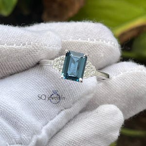 May include: A silver ring featuring a rectangular, teal-colored gemstone as the centerpiece. The ring is flanked by smaller, clear stones on each side. The ring is held against a white fabric background. The text "SQ jewels In" is visible.