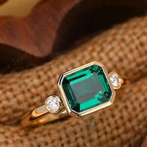 Könnte beinhalten: Ein goldener Ring mit einem großen, smaragdgeschliffenen grünen Edelstein, flankiert von zwei kleineren, runden, klaren Edelsteinen. Der Ring hat ein poliertes Goldband und eine Fassung, die das leuchtende Grün des zentralen Steins hervorhebt.