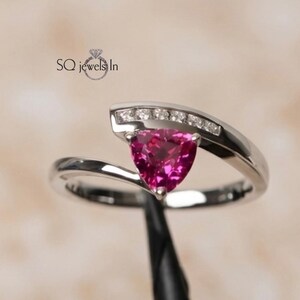 May include: A silver-toned bypass ring featuring a trillion-cut vibrant pink centre stone and a curved channel set with small clear accent stones. The text SQ jewels In is visible above the ring. The design has a polished metallic finish.