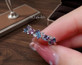 Puede incluir: Un anillo de plata con un grupo de piedras preciosas en tonos azules, morados y transparentes. El anillo tiene un diseño único con piedras rectangulares y redondas. El anillo lo sostiene una persona.