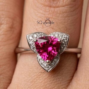 May include: A silver-toned ring featuring a large, triangular, vibrant pink gemstone at the center. The stone is surrounded by a halo of small, clear, sparkling stones set in a petal-like frame. The text SQ jewels In is visible above the ring.