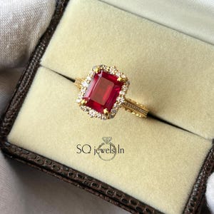 Può includere: Un anello d'oro con una grande gemma rossa rettangolare, circondata da pietre bianche più piccole. L'anello è presentato in una scatola con la scritta "SQ jewels In". La fascia dell'anello ha un design dettagliato.