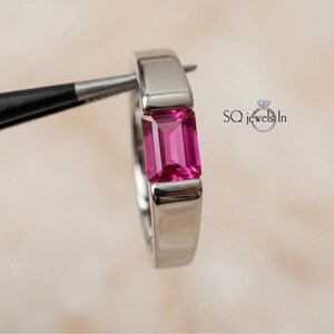 May include: A silver-toned metal ring featuring a single emerald-cut vibrant pink gemstone set in a smooth bezel setting. The ring is held by black tweezers and includes the text SQ jewels In with a small diamond icon printed on the right side.