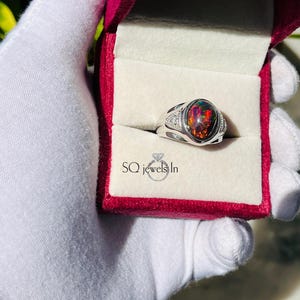 Könnte beinhalten: Ein silberner Ring mit einem großen, ovalen Opal-Edelstein. Der Opal zeigt eine lebendige Farbpalette, darunter Rot, Orange und Schwarz. Kleine, klare Steine sind auf beiden Seiten des Opals gesetzt. Der Ring wird in einer roten Schmuckschatulle präsentiert.