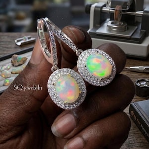 May include: A pair of silver earrings with oval opal gemstones. Each opal is surrounded by a halo of small, clear gemstones. The opals display a play of color, with hints of green, yellow, and red. The earrings are held in a hand.