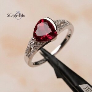 May include: A silver-toned ring featuring a large, trillion-cut deep red gemstone as the centerpiece. The band is split into two arms, each accented with small, clear round stones. The text SQ jewels In is visible in the upper left corner of the image.