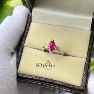 May include: A silver ring with a marquise-cut, vibrant red gemstone as the centerpiece. The ring is set with smaller, clear gemstones on either side of the main stone. The ring is displayed in a brown jewelry box with a beige interior. The text "SQ jewels In" is at the bottom.