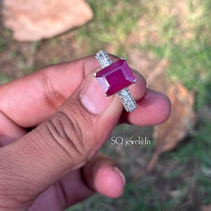 Peut inclure: Une bague en argent ornée d'une grosse pierre rouge foncé de forme rectangulaire. La monture est ornée de plus petites pierres précieuses transparentes. La bague est tenue sur un fond flou vert et marron. Le texte "SQ jewels In" est visible.