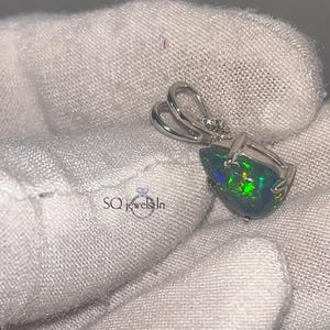 以下が含まれることがあります： 黒オパール宝石をあしらった、洋梨型のペンダント。緑と青の虹色の色合いが特徴です。ペンダントはシルバーの金属フレームにセットされ、チェーン用のループが付いています。「SQ jewels In」の文字が含まれています。