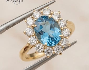 May include: A gold ring featuring a large, oval-cut, light blue gemstone surrounded by a halo of smaller, round, white gemstones. The ring is held by tweezers, showcasing the intricate design and craftsmanship of the jewelry.