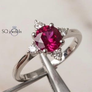 May include: A silver-toned ring featuring a large, round, vibrant red center stone held by prongs, accented by four smaller, clear, round stones arranged in a cross pattern. The text SQ jewels In is visible on the left side of the ring.
