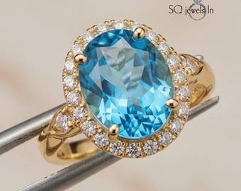 May include: A gold ring featuring a large, oval-cut, light blue gemstone surrounded by a halo of smaller, clear gemstones. The ring's band has intricate detailing. The text "SQ jewels In" is visible.