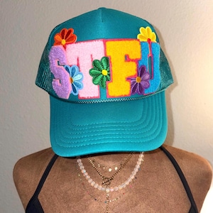Puede incluir: Gorra de camionero turquesa con las letras "STFU" en rosa, amarillo y azul, decorada con flores de fieltro de colores. La gorra tiene una parte trasera de malla y una visera curva. Un accesorio divertido y llamativo.