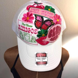 Puede incluir: Gorra de camionero blanca con una parte trasera de malla y una visera curva. La parte delantera presenta parches bordados de flores, una mariposa y el texto "Save water drink margs" y "The Three AMIGOS".
