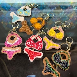 May include: A collection of beaded bikini and underwear keychains in various colors, including blue, yellow, pink, and red. Each keychain features a gold-colored ring and chain.