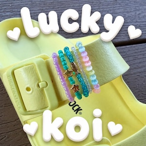 May include: Five beaded bracelets resting on a light yellow Birkenstock sandal. The bracelets feature purple, teal, clear, and multicolored beads, with gold-toned star and fish charms. The text "lucky koi" is overlaid in white bubble letters.