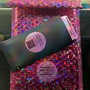 K&ouml;nnte beinhalten: Ein metallisch pinker Luftpolsterumschlag enth&auml;lt einen schwarzen Umschlag mit einem holografischen runden Aufkleber mit dem Text Sparkle Parcel Co. Unique Handmade Crafts und einem QR-Code. Eine Dankeskarte steckt oben im Umschlag.