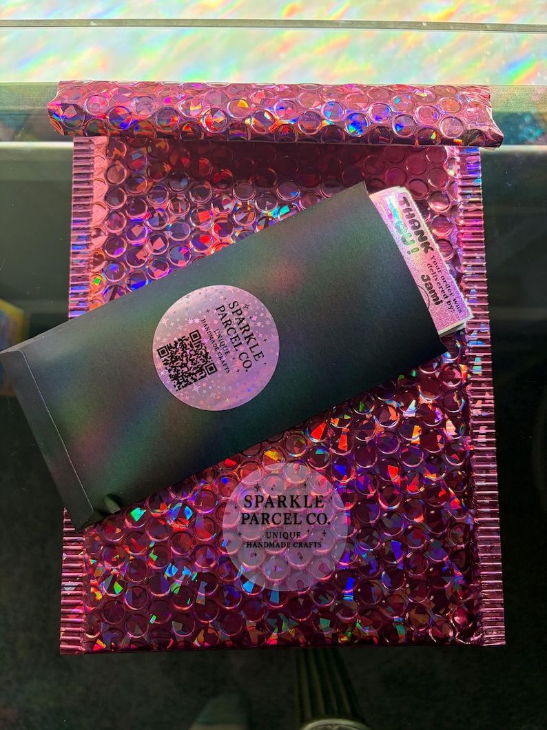 K&ouml;nnte beinhalten: Ein metallisch pinker Luftpolsterumschlag enth&auml;lt einen schwarzen Umschlag mit einem holografischen runden Aufkleber mit dem Text Sparkle Parcel Co. Unique Handmade Crafts und einem QR-Code. Eine Dankeskarte steckt oben im Umschlag.