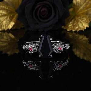 Peut inclure: Bague en argent avec une grande pierre noire en forme de cercueil en son centre. La bague présente des accents de roses et de pierres précieuses rouges sur le bracelet. La bague est posée sur un fond sombre avec une rose noire et des feuilles dorées.