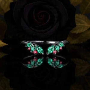 Peut inclure: Une bague en argent au design unique, ornée de grappes de pierres précieuses vert émeraude et rouge rubis. La bague est posée sur un fond sombre avec une rose noire et des feuilles dorées, créant un contraste saisissant. Les pierres précieuses se reflètent.