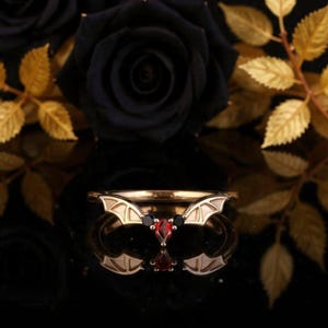 Peut inclure: Une bague dorée ornée d'ailes de chauve-souris et de pierres précieuses rouges et noires. La bague a une esthétique gothique. Le fond présente des roses noires et des feuilles dorées.