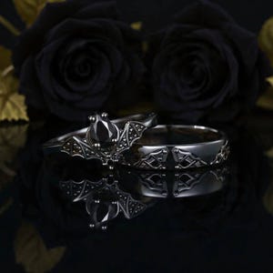 Peut inclure: Deux bagues noires sur le thème gothique. L'une présente un motif de chauve-souris avec une pierre noire, l'autre un motif gravé. Les bagues sont présentées sur une surface réfléchissante, avec des roses noires en arrière-plan.