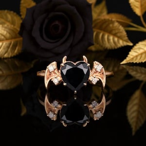 Puede incluir: Un anillo dorado con una piedra preciosa negra en forma de corazón, acentuado con pequeñas piedras preciosas blancas y detalles de alas. El anillo está sobre un fondo negro con una rosa negra y hojas doradas.