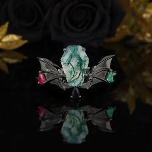May include: A unique ring featuring a large, coffin-shaped, green and white gemstone as the centerpiece. The ring has a dark metal setting with bat wing accents and smaller red and green gemstones on either side. A dark, reflective surface enhances the image.