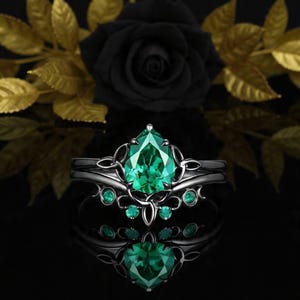 Peut inclure: Un ensemble de bagues noires avec une grosse pierre verte émeraude en forme de poire au centre. La bague est ornée de petites pierres vertes et de détails de filigrane complexes. Le fond présente une rose noire et des feuilles dorées.