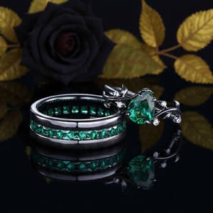 Peut inclure: Deux bagues en métal sombre avec des pierres précieuses vert émeraude. L'une présente une rangée d'émeraudes carrées, l'autre une émeraude en forme de poire et des motifs floraux avec de petites pierres blanches.