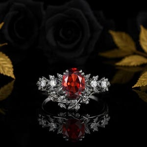 Peut inclure: Une bague en argent ornée d'une pierre précieuse ovale rouge foncé au centre. La bague est ornée de petites pierres blanches et de détails en forme de feuilles. La bague est posée sur un fond noir avec un reflet.