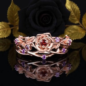 Peut inclure: Bague en or rose avec un motif de rose, une pierre centrale rouge et des pierres d'accent violettes. La bague présente un design floral et en forme de vigne, avec une finition polie. La bague est présentée sur une surface réfléchissante.