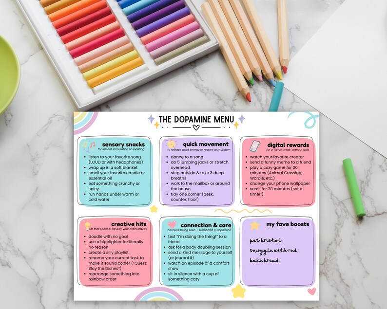 May include: A colorful print titled "The Dopamine Menu" with a pastel rainbow design. The menu lists sensory snacks, quick movement, digital rewards, creative hits, connection & care, and favorite boosts. Colored pencils and pastels are in the background.