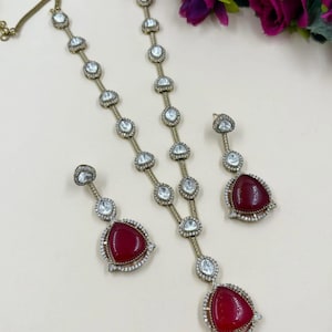 May include: A jewelry set featuring a necklace and earrings. The necklace and earrings have a gold-tone chain with clear, round stones. The earrings and necklace pendants have a large, triangular, deep red stone surrounded by smaller clear stones.