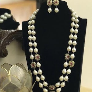 Op de afbeelding: Een parelketting met twee strengen en bijpassende oorbellen. De ketting heeft grote, druppelvormige parels met sierlijke goudkleurige accenten en rode edelstenen. De oorbellen hebben een vergelijkbaar ontwerp, met een rode edelsteen in het midden en een bungelende parel.