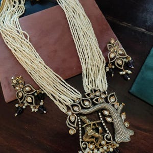 May include: An elaborate necklace and earring set. The necklace has a tree-shaped pendant with a gold figure playing a flute, and is made of multiple strands of small, white beads. The earrings have a similar design with black and white details.