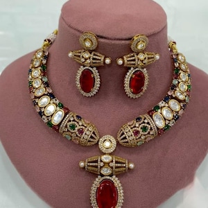 May include: An elaborate gold necklace and earring set. The necklace and earrings are adorned with a variety of colourful gemstones and clear crystals. The central feature of the necklace and earrings is a large, oval-shaped, red gemstone. The jewellery is displayed on a pink mannequin head.