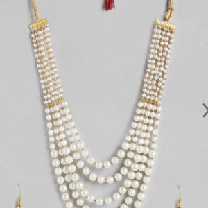 May include: A multi-strand pearl necklace with matching earrings. The necklace features multiple strands of white pearls, connected by gold-coloured accents. A red tassel hangs from the top. The earrings each have a single pearl.